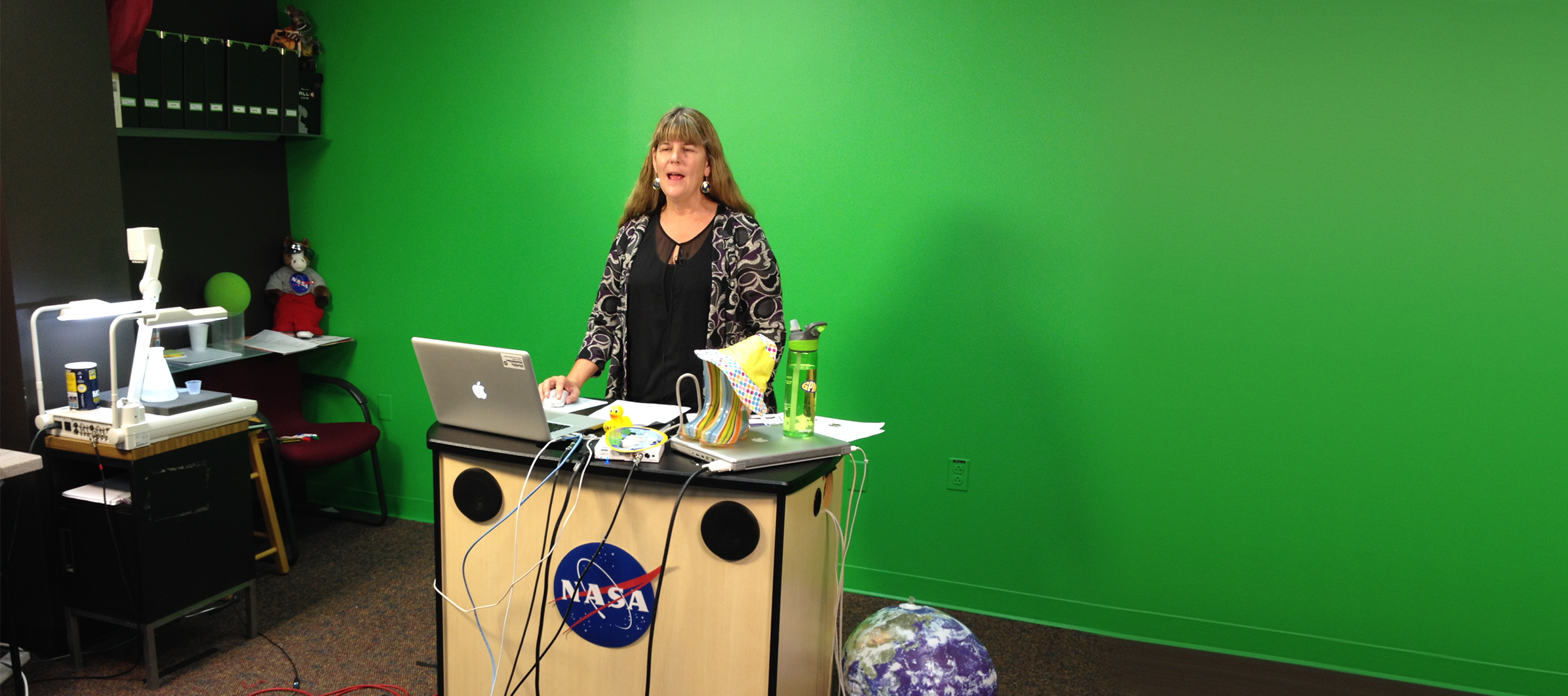 A woman stands giving a presentation at a desk in a studio, in front of a green screen wall.